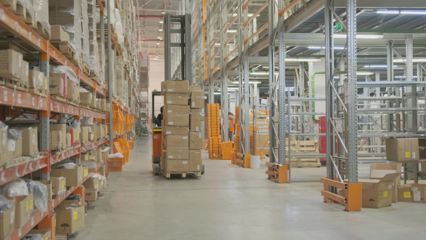Worker in warehouse rides forklift moving pallets of cardboard boxes of goods between row of racks. Royalty-Free Stock Footage #1076500040