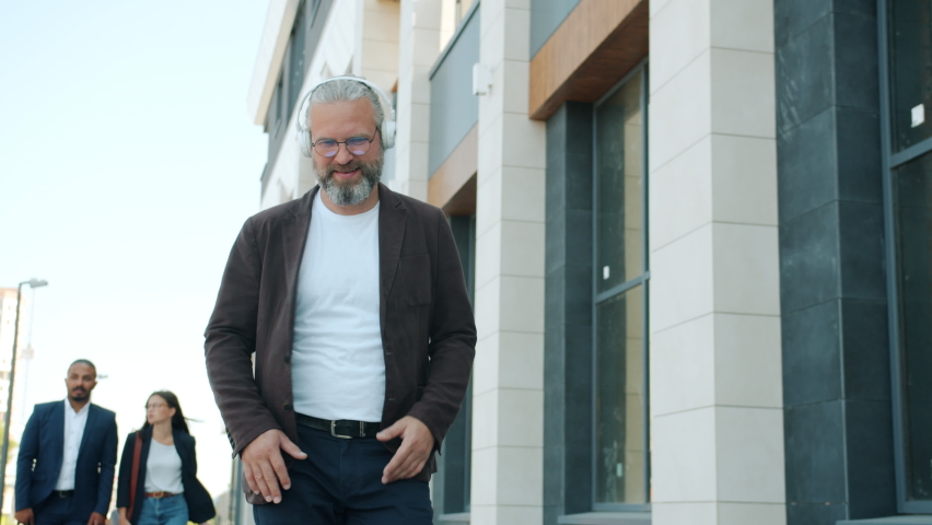 Careless mature man is walking outdoors wearing headphones dancing enjoying music in urban street. Happy business people and modern technology concept.