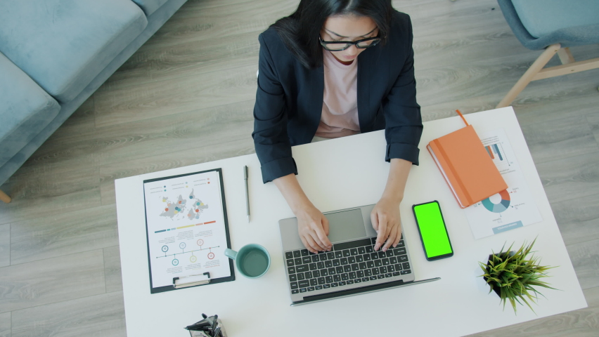High angle view of young Asian businesswoman working at home using laptop computer and looking at chroma key green screen smartphone at desk