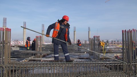 OMSK, RUSSIA - FEBRUARY 04 2021: Happy employee in orange vest takes metal rod working with colleagues at construction site of sports stadium on winter day on February 04 in Omsk
