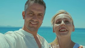 Young woman in white dress and her husband happily video call with their loved ones on their mobile phones with sea view in the background. Happy couple enjoying their vacation.Front camera view.
