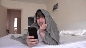 A Handsome Man Lies On A Bed Under A Blanket In His Bedroom And Flips A Tape On His Phone. Read News in Smartphone, Networking