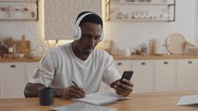 Young african student having video chat on modern smartphone while staying at home. Male student in wireless headphones enjoying distance learning.