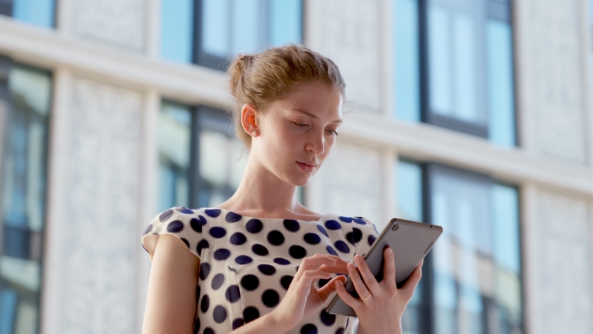 Medium shot of beautiful businesswoman with digital tablet outdoor. Low angle view of young female manager with tablet pc looking and smiling at camera outside office building