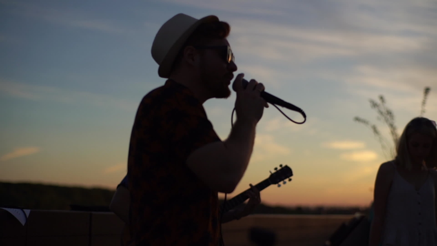 Silhouette of young talented music band performing in city embankment during sunset. Stylish bearded singer and guitarist give performance for public in city park. | Shutterstock HD Video #1076541107