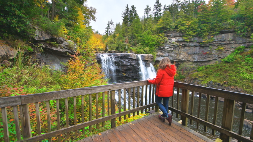 Young woman standing leaning on wooden boardwalk railing at Blackwater Falls waterfall state park, West Virginia at colorful autumn fall season with water flowing