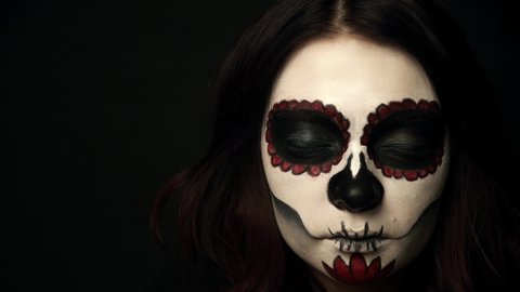 Lady with sugar skull makeup opens green eyes and her pupils get smaller. Portrait of woman on black background watching in camera. Theme of santa muerte makeup and emotions.