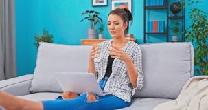 Young female employee sits on couch with a laptop on lap and takes a call from client, conducts conversation over internet, explains issue, clarifies, accepts complaint, discusses terms and conditions