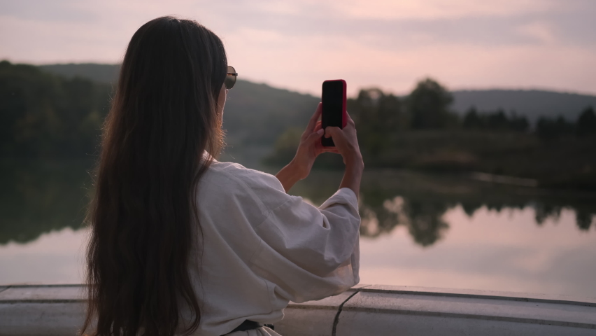 Young brunette with mobile taking picture of nature scene with quiet river and woods. Adding new photos to landscapes collection