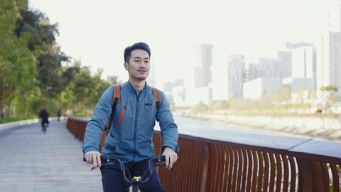 young asian man riding bicycle on riverfront path looking at view
