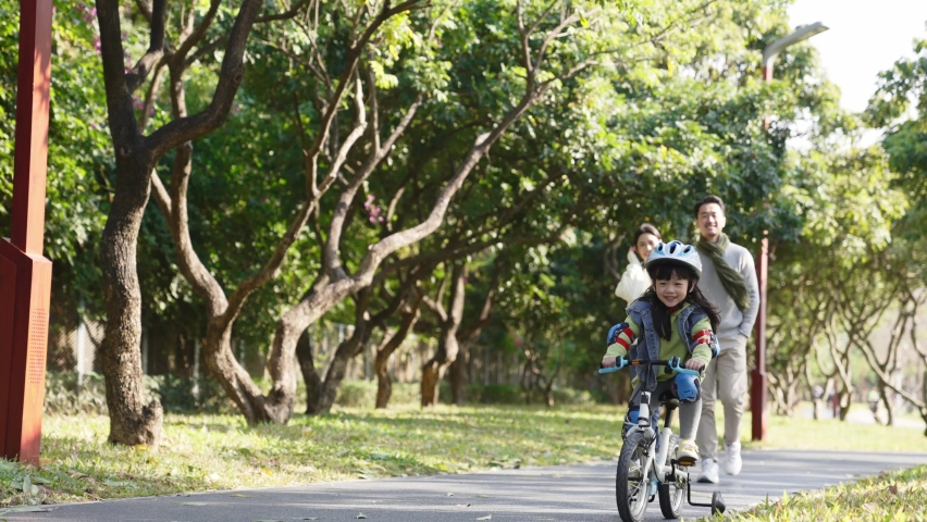 Little asian girl with full protective gears riding bike in park while parents watching from hehind | Shutterstock HD Video #1076558129