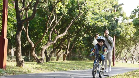 little asian girl with full protective gears riding bike in park while parents watching from hehind