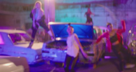 Group of stylish young people dancing , moving inside abandoned garage building . Old hippy style party . Colorful stylish clothes , african braids . Dancers on the car hood , vintage broken old cars 