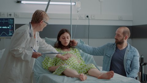 Obstetrician giving advice to young pregnant woman and husband sitting in hospital ward bed. Caucasian family with pregnancy getting parenthood support from childbirth doctor at maternity