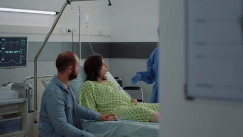 Caucasian couple expecting baby in maternity ward at hospital. Pregnant woman sitting in bed talking to african american nurse and young husband. Medical assistance for childbirth
