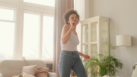 Happy african american woman dancing jumping at home, having fun celebrating with funny dance. Singing karaoke on the hairbrush like microphone and fooling around, music concept.