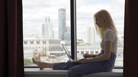 Young joyful woman talking on a video call using a laptop while sitting on a windowsill by a large window overlooking the skyscrapers. Living in a big city, communication at a distance.