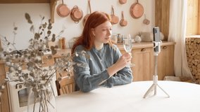 Happy red haired beautiful teen girl celebrating holding glass of champagne on front of cell phone. Having video call chat long distance relationship video. Blue shirt. Sunny interior rustic style