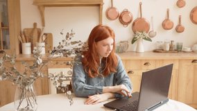 Beautiful red haired teen girl using laptop sitting at kitchen table having online meeting conversation. Chatting online video call. Remote work video footage. Blue shirt. Sunny interior rustic style