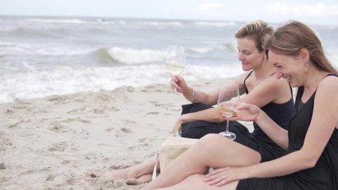 Two beautiful women sit on a sandy beach with glasses of wine. Same-sex love, a date by the ocean
