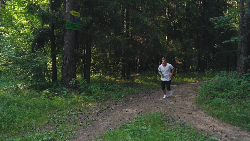 Adult fit Caucasian male is jogging on summer forest trail, he is stopped to drink water. 4K