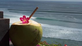 Video footage of green young fresh coconut close up with bamboo straw and pink tropical flower on the  cliff and blue ocean with big waves for surfing on background in Bali