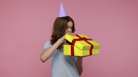 Bad present. Attractive teenager girl in blue T-shirt carefully unpacking gift, looking inside box and expressing disappointment, looks unhappy. Indoor studio shot isolated over pink background.