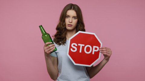 Stop Drinking. Serious teenager girl wearing casual attire holding stop sign and alcohol bottle, looking at camera with serious expression, warning. Indoor studio shot isolated over pink background.