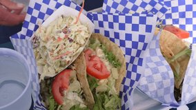 This delicious video shows carry out baskets filled with fish tacos and coleslaw at an outdoor restaurant with paper blowing in the wind.