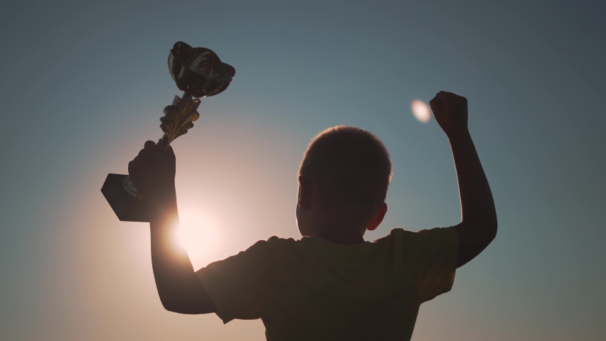 Winner child silhouette. Kid with cup of success raises her hand. Silhouette of child at sunset. Winner success. Victory, path to dream. Child won the victory. Cup in hand of a kid. Superhero kid