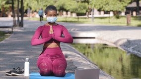 Black instructor holding online lessons, shows breath technique to reach inner harmony, stress relieve sitting on sport mat outdoor. Young lady in sportswear and protective medical mask using laptop