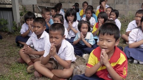 Yala, Thailand July 8, 2016: Thai group of young children girls and boys together praying, closed eyes, chanting praying hands, sitting on the ground. Primary students Thetsaban school