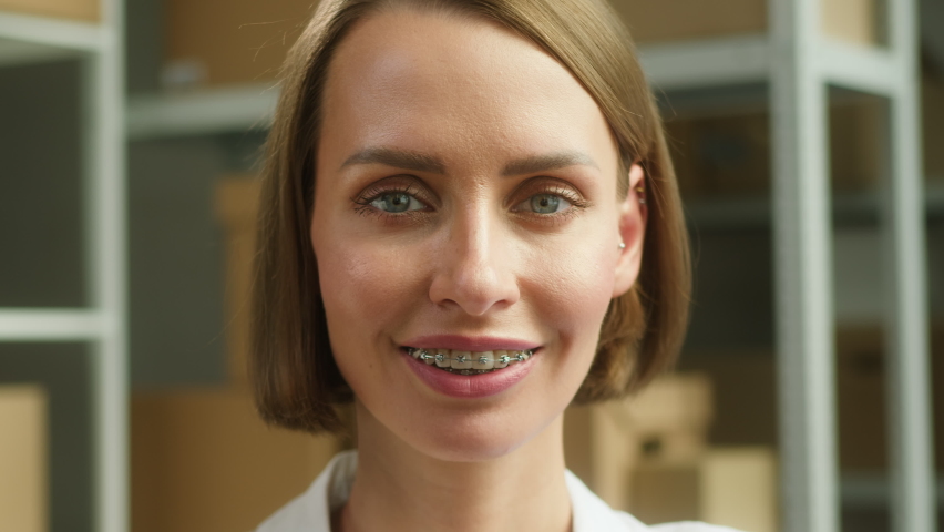 Young smiling woman sitting in storage close-up, smile with braces of female employee of warehouse, looking in camera on background of cardboard boxes. Delivering and logistics, small woman business.