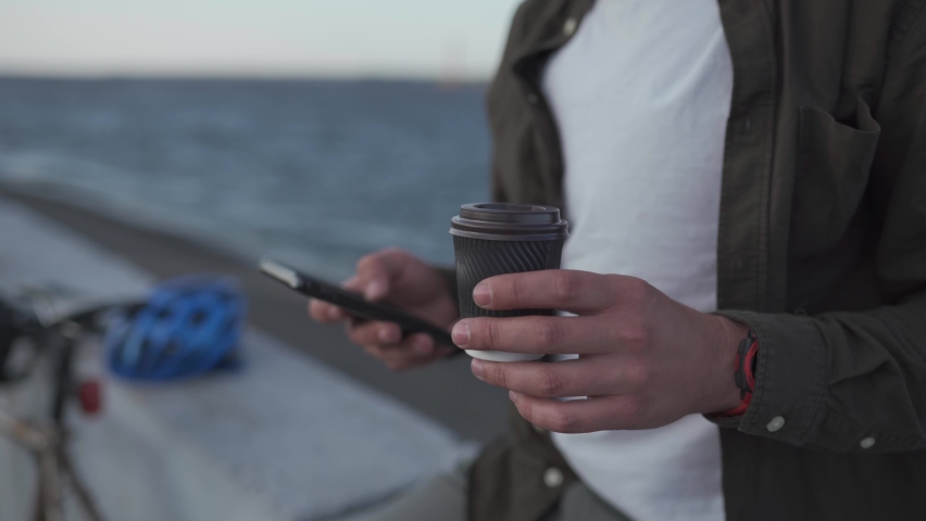 Reading news, social networks, surfing Internet. 5G on your smartphone. Coffee break. Close-up of male cyclist hand holding smart phone while drinking takeaway coffee near bike on promenade by sea