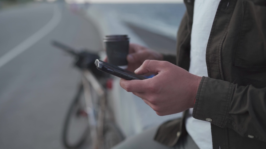 Unrecognizable male cyclist use smartphone and drinks takeaway coffee while standing near bike on embankment. Delivery man on bike during coffee break on the bike path by sea. Social networks outdoor