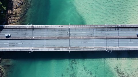 Busy coastal road and pedestrian bridge spanning a clear ocean estuary allowing people and vehicles to cross. Drone view looking down