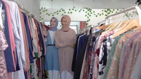 asian veiled waitresses while serving female shoppers choosing clothes on hangers in boutique shops