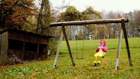 Halloween. Evil clown  in a bright fancy dress is spinning on a swing. Slow motion. Horror and fear