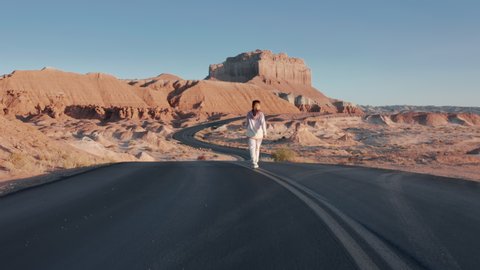 Back view of woman in casual bright sweatshirt and joggers walking by winding road with red mountains nature landscape. Relaxed woman traveling famous landmark Goblin Valley park. Utah aerial 4K, USA