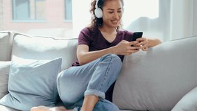 Video of smiling young woman listening to music with smartphone while sitting on sofa at home.