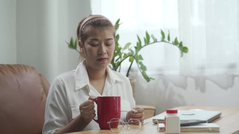 Young Asian Woman feeling sick ,Taking a Pill and Drinking a Glass of Water Sitting on Couch at Home.