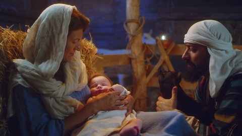 Joseph stroking lamb and speaking with Mary embracing sleeping baby Jesus near stalls in dim barn in Bethlehem nativity scene of Christmas