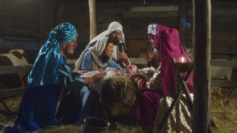 Zoom in view of Magi and parents gathering around manger with baby Jesus and talking in inn stable in Bethlehem