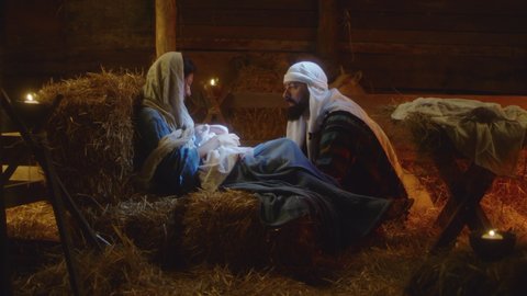 Zoom in view of Joseph talking with Mother Mary caressing baby Jesus on Christmas day in dark stable in Bethlehem