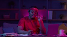 African American Male Hotline Operator Talking With Customer Wearing Headset Sitting At Laptop Computer Working Indoor At Night. Man Communicating With Clients In Helpline Office. Low Light