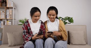 Happy asian twin girls playing game on smartphone together while sitting on couch at living room. Funny women playing game on mobile phone. Hobby, techology and lifestyle concept.