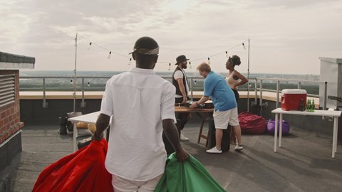 Handheld tracking shot of African-American man carrying bean bags and showing up to rooftop party, then hugging friends and shaking their hands