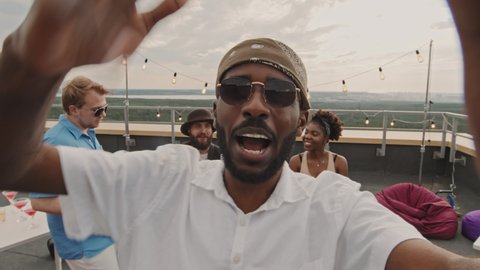 POV shot of happy African-American man holding camera and singing while filming himself at party with friends on rooftop terrace
