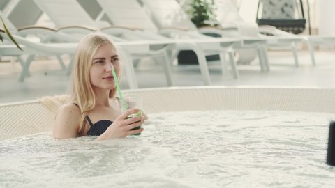 Charming blonde woman resting in jacuzzi with soft drink. Spending vacations at the resort.