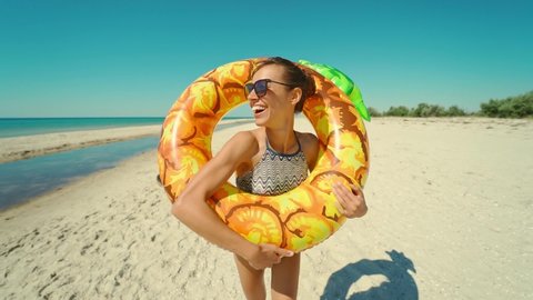 Cheerful beautiful young millennial woman running on empty sunny sandy beach holding yellow inflatable pineapple float in summer.